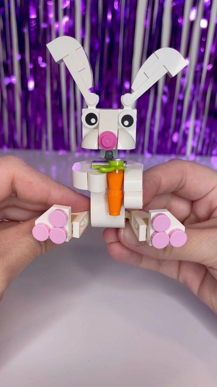 White LEGO bunny figure holding an orange carrot held by two hands.