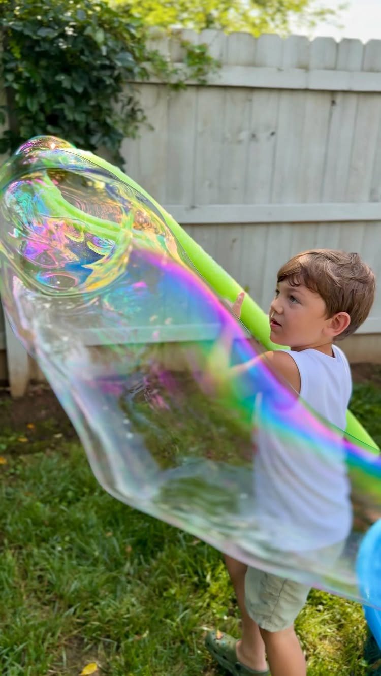 Young boy creating a giant iridescent soap bubble in a grassy backyard