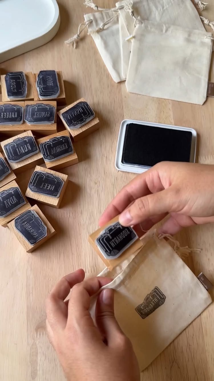 Hands stamping a small beige fabric bag using wooden rubber stamps and ink.