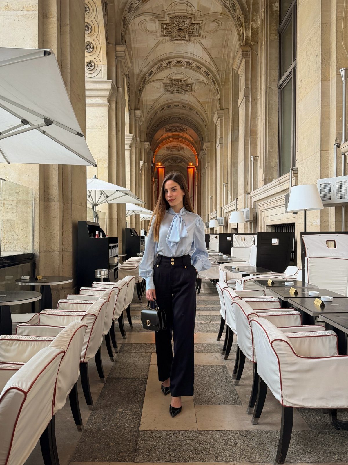 Woman in light blue shirt and black trousers standing in a vaulted arcade.