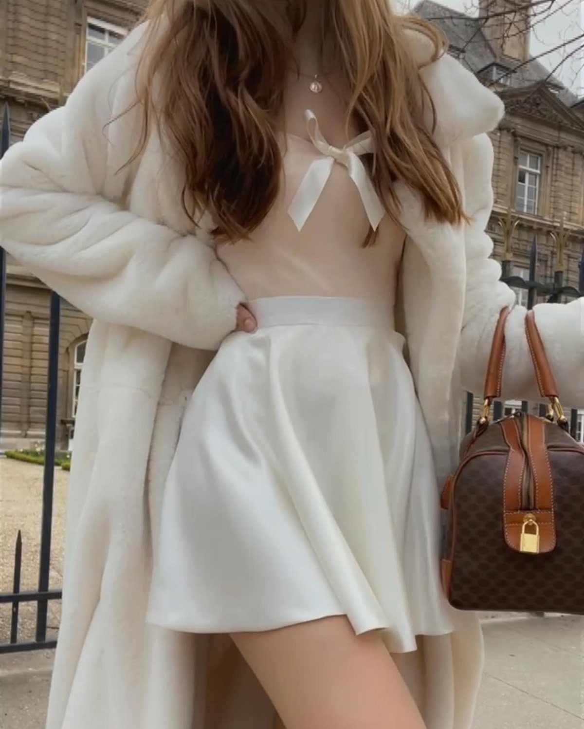 Woman in white faux-fur coat and cream satin skirt holding a brown handbag.