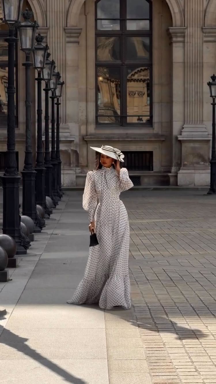 Woman in a polka dot maxi dress and hat holding a black clutch bag.