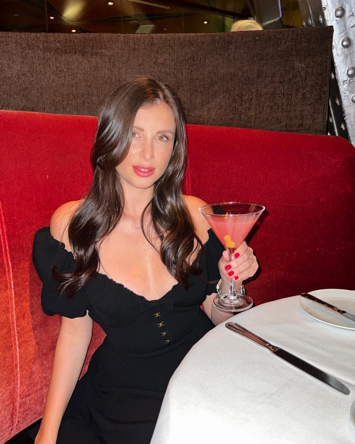Woman in black off-the-shoulder dress holding a pink cocktail in a red booth.