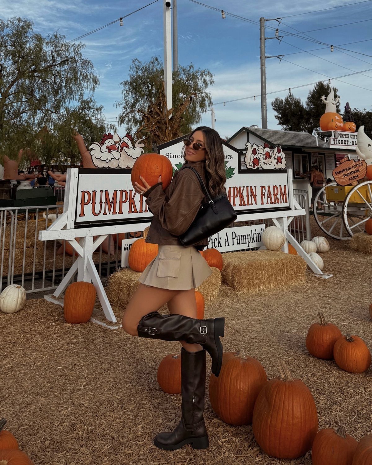 Woman in brown jacket and knee-high boots holding a pumpkin at a farm.