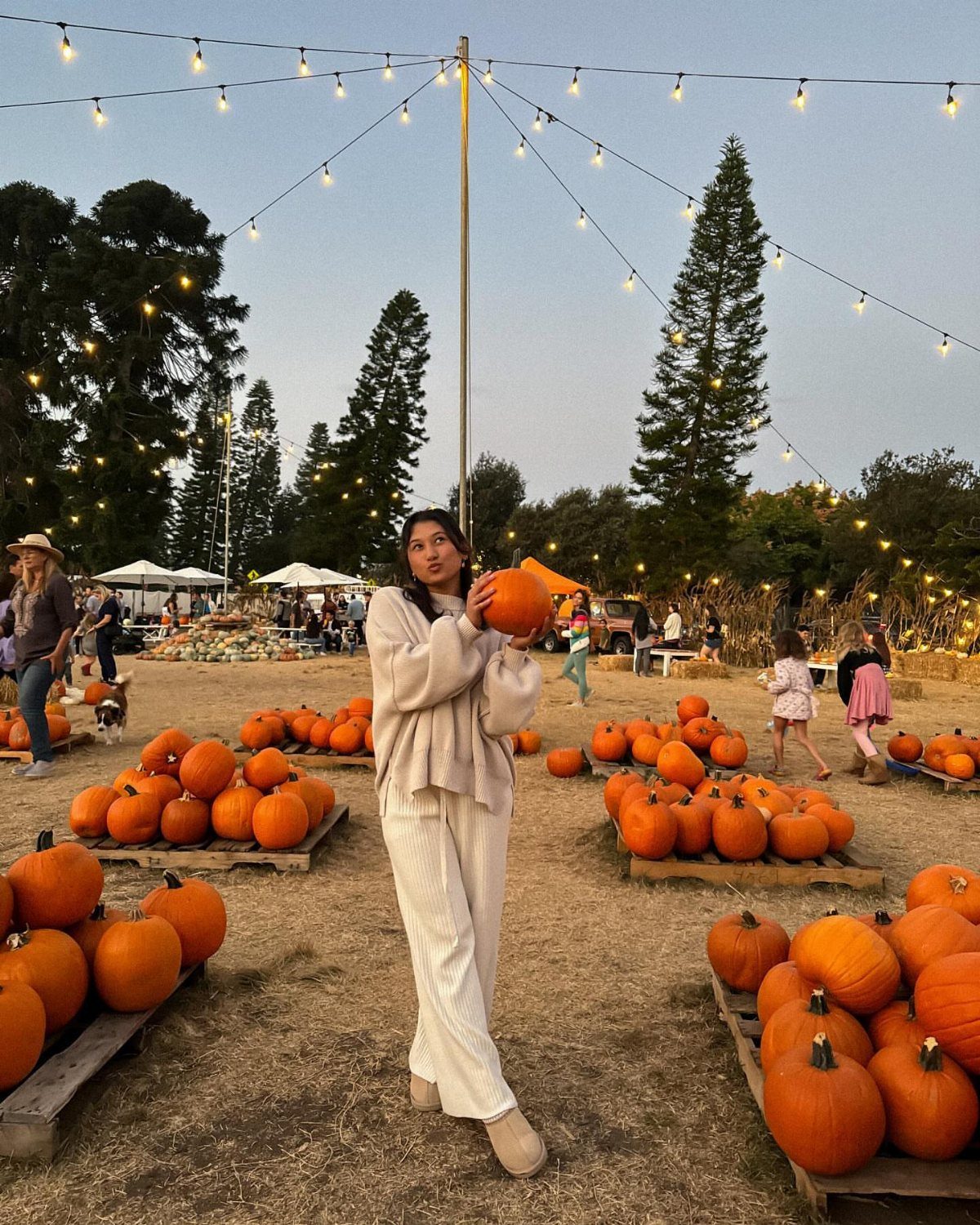 Woman in cream sweater and trousers holding a pumpkin in a pumpkin patch.