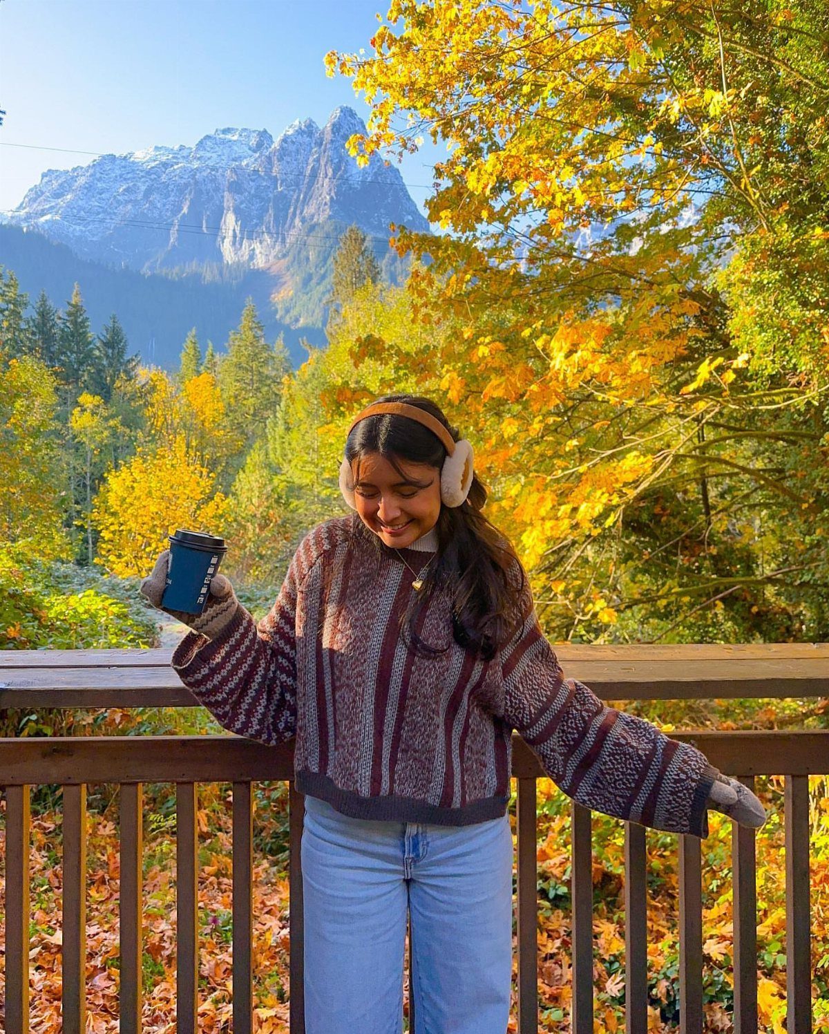 Woman in patterned sweater and blue jeans holding coffee against autumn mountain backdrop