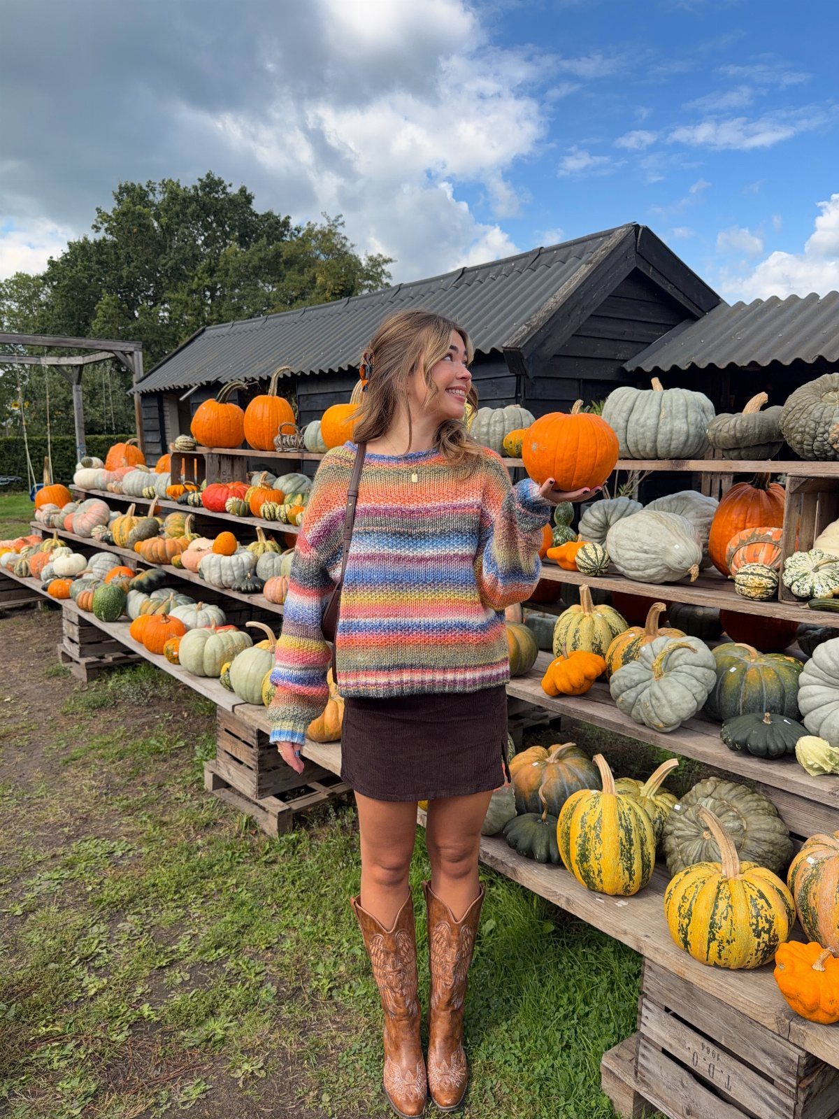 Woman in striped sweater and cowboy boots holding a pumpkin at a patch.