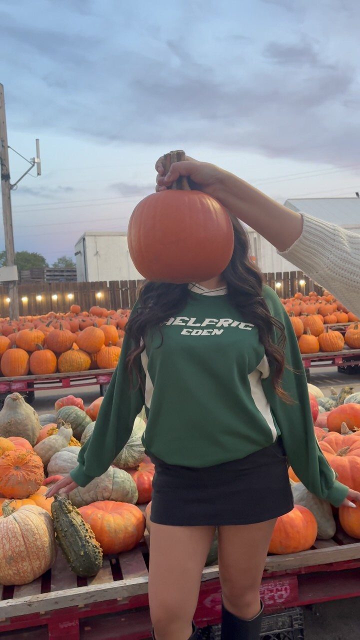 Person in green sweatshirt and black skirt standing among many orange pumpkins