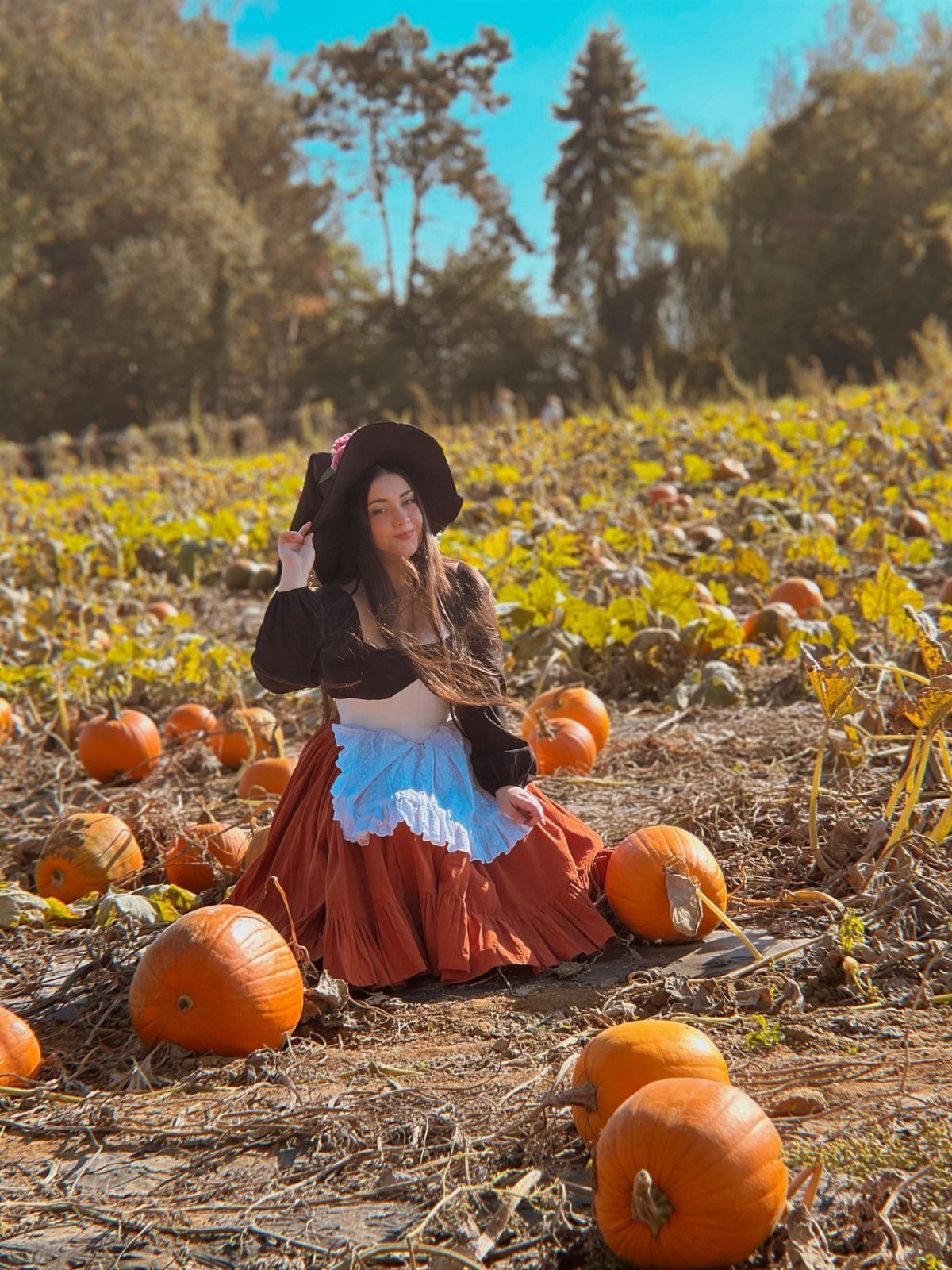 Woman in brown witch hat and orange skirt sitting in a pumpkin patch