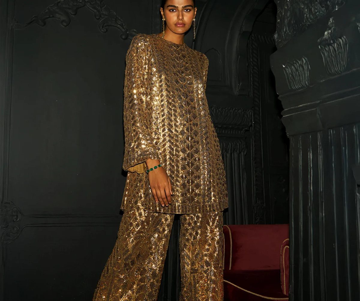 Woman in a gold sequined tunic and matching wide-leg trousers in a dark room.