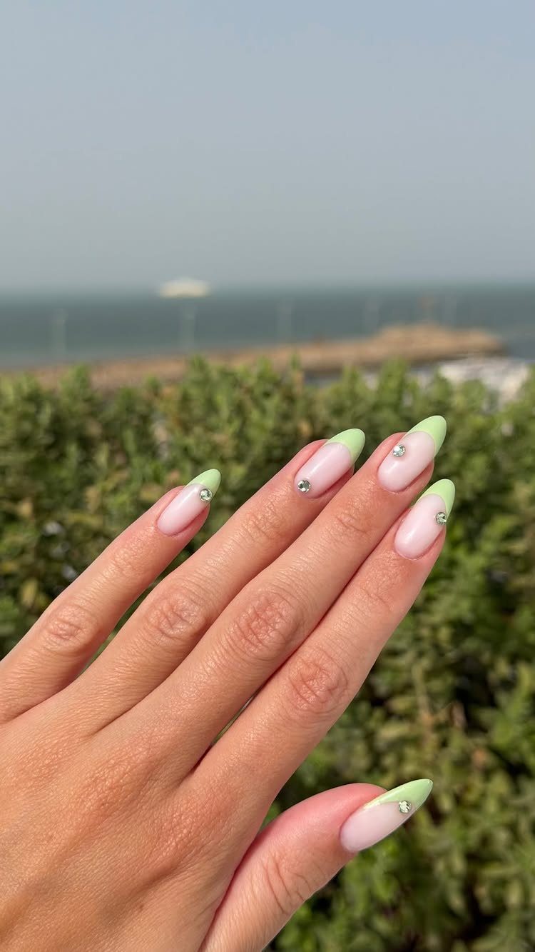 Almond-shaped nails with light pink bases, bright green tips, and silver rhinestone accents.