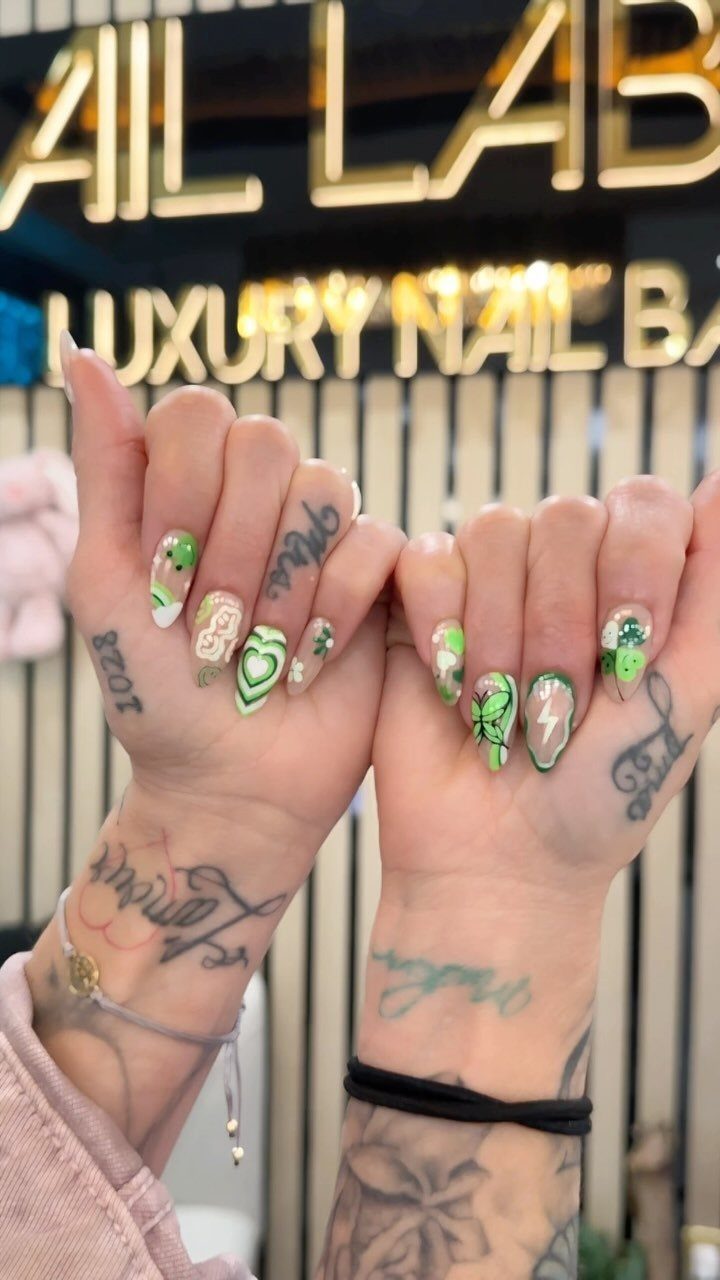 Green and gold heart nails with shamrock accents on tattooed hands.