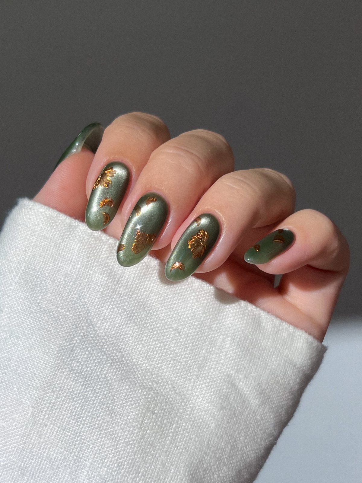 Olive green metallic nails with gold foil flakes on a hand wearing white.