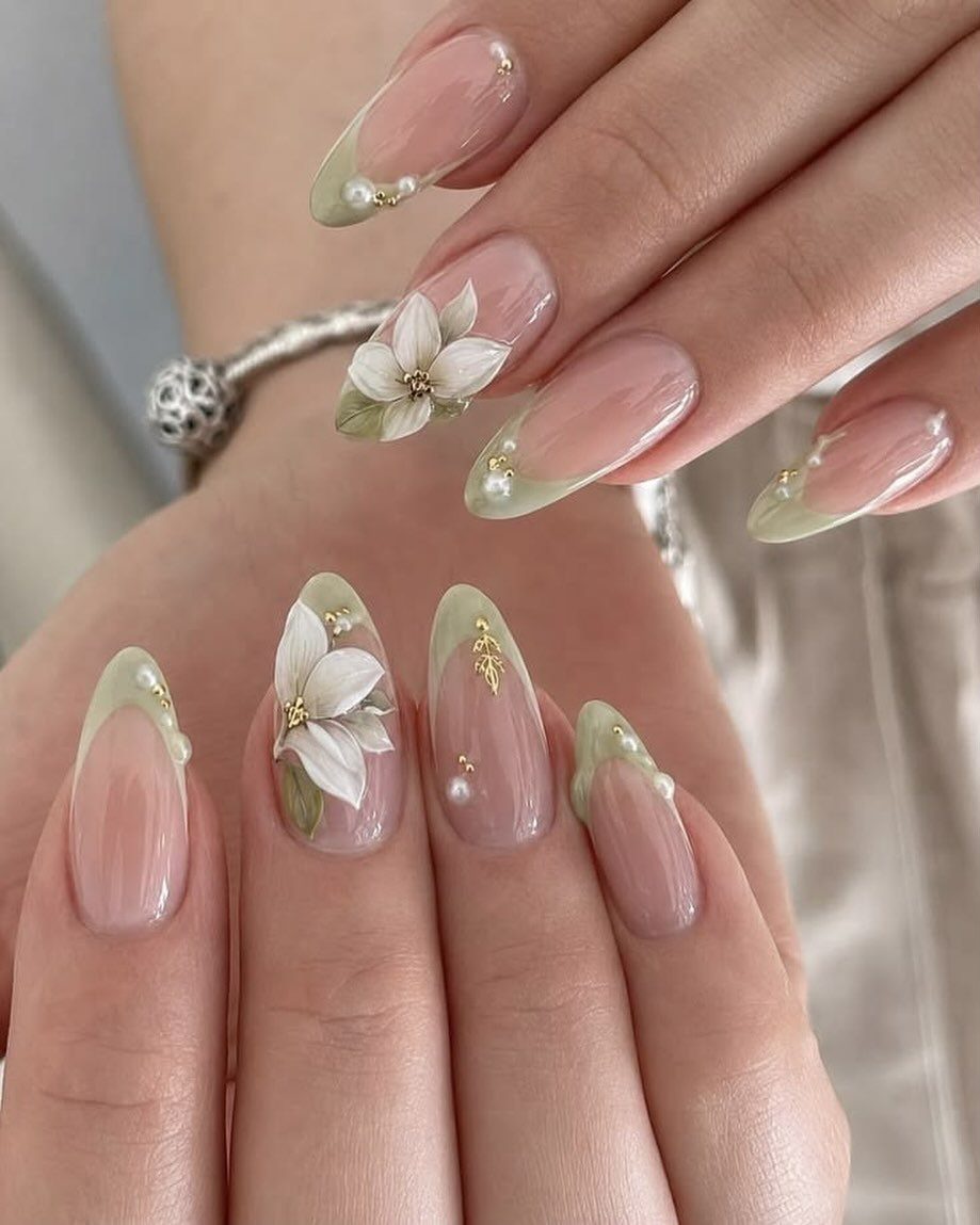 Almond nails with nude base, pastel green tips, white flowers and gold accents.