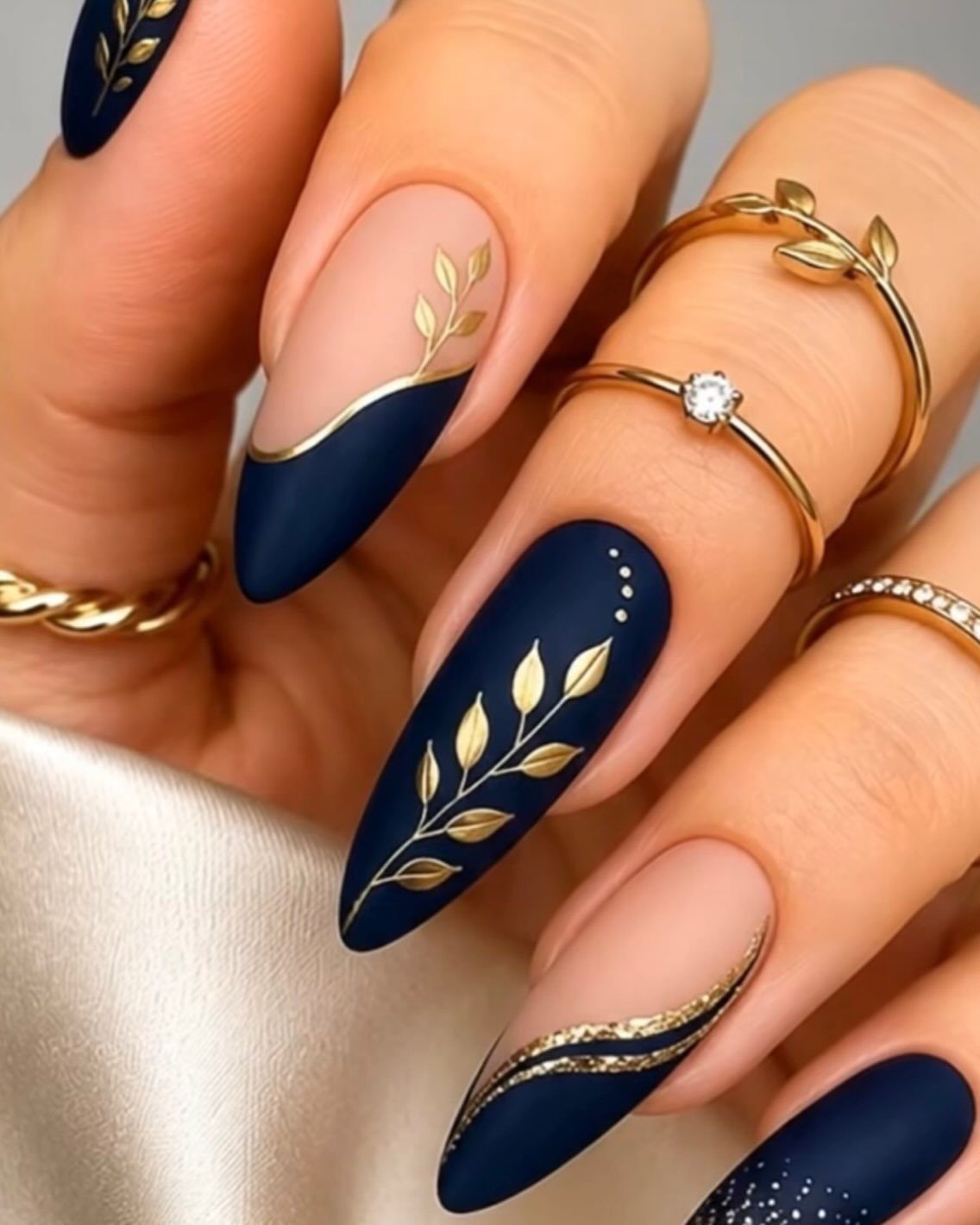 Navy blue and nude almond nails with gold leaf and glitter details.