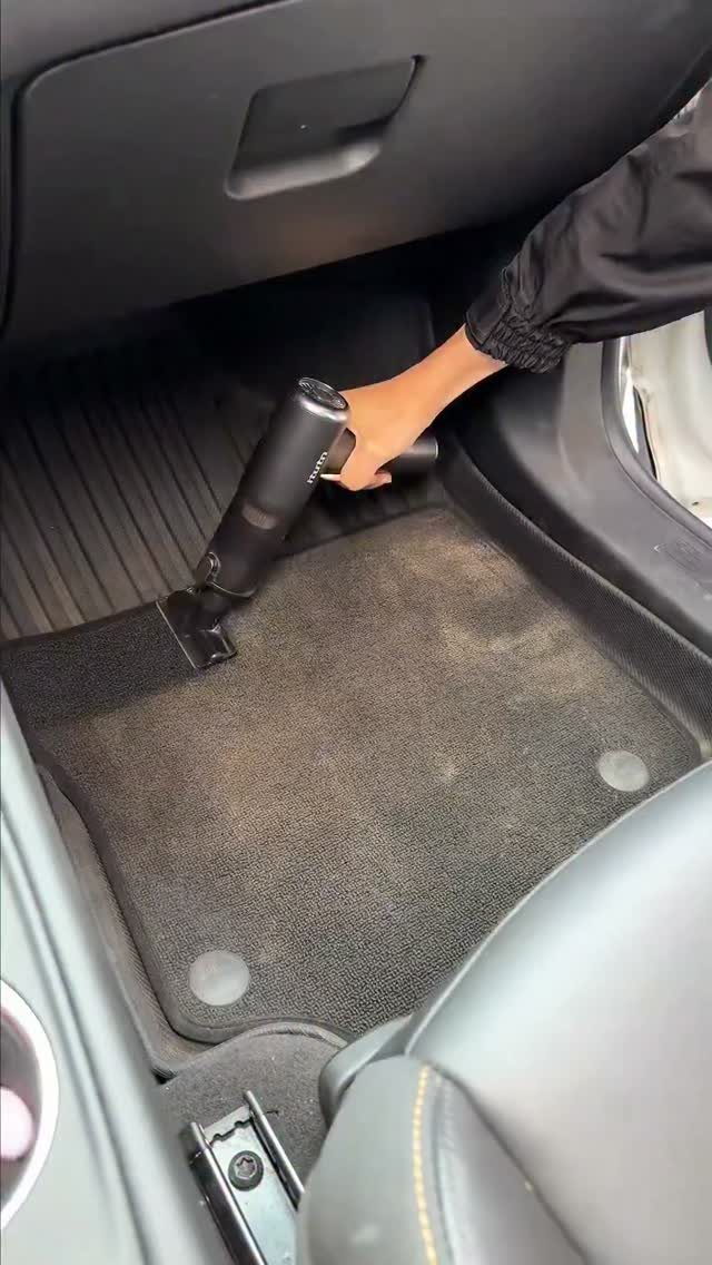 Handheld cordless vacuum cleaner cleaning a grey car floor mat inside a vehicle.