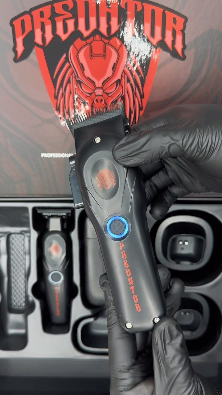 Black electric hair clipper held by gloved hands against a Predator themed box.