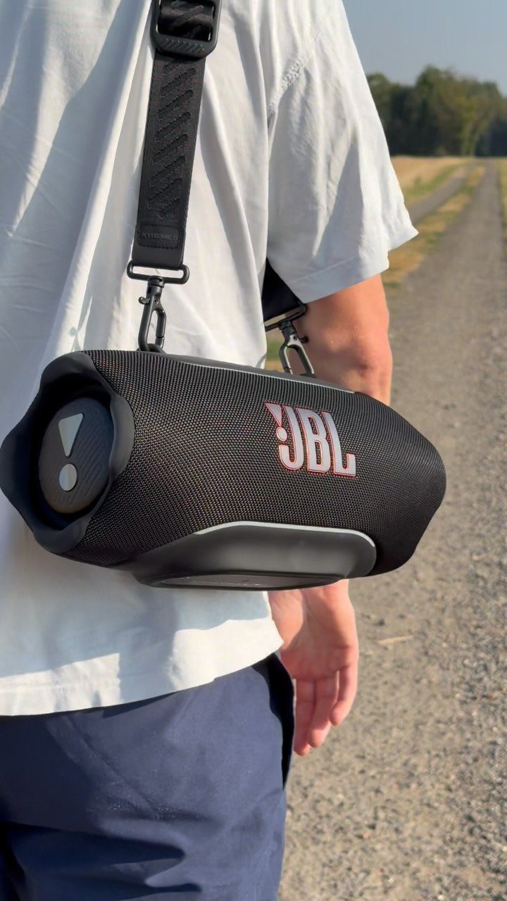 Black JBL portable Bluetooth speaker carried by a person on a gravel path