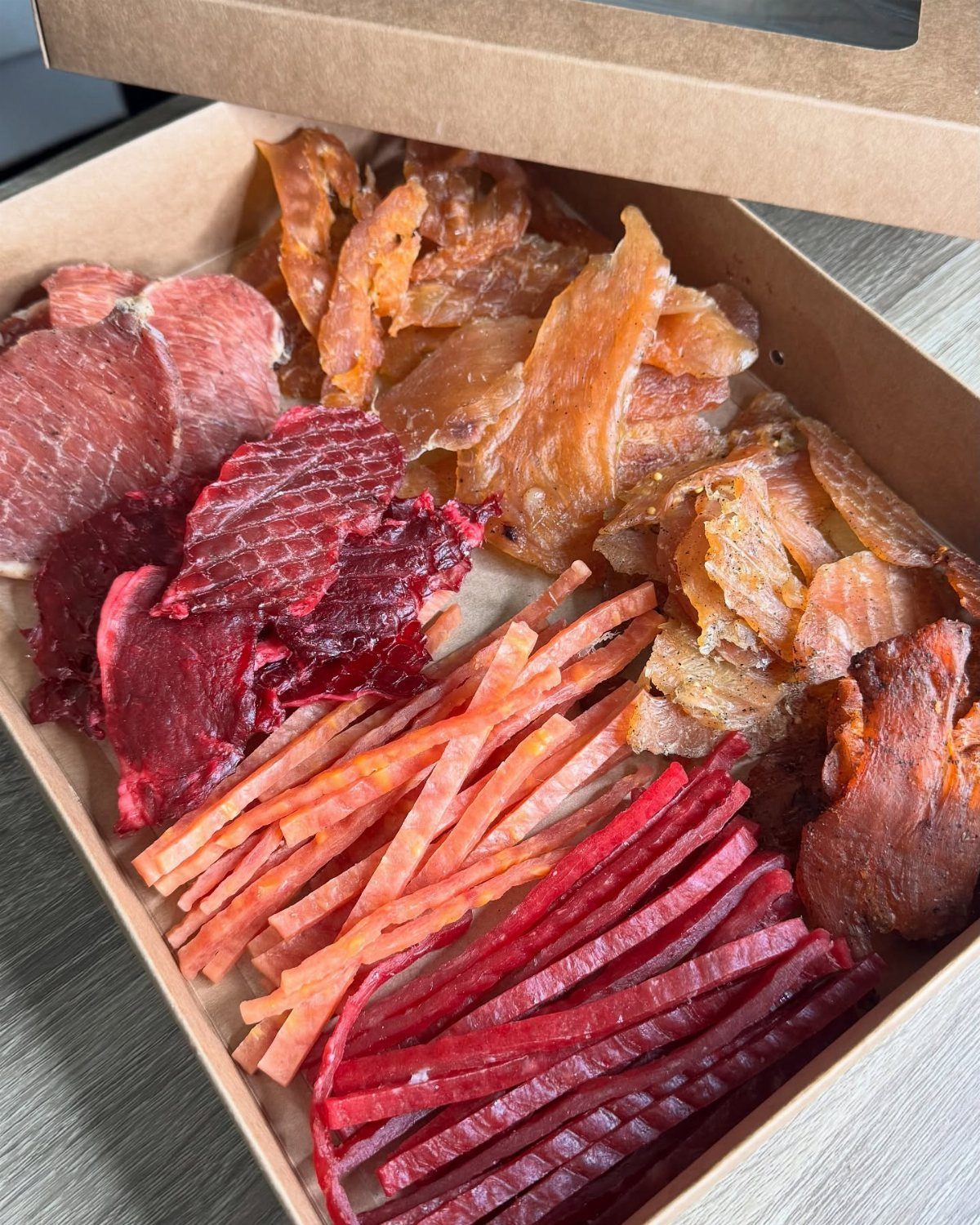 Assorted dried meat jerky strips and slices arranged inside a brown cardboard box.