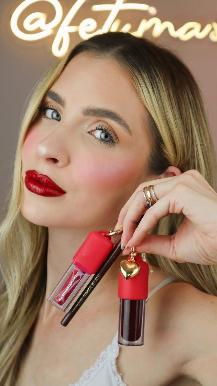 Woman with bold red lipstick holding liquid lipstick tubes and a makeup brush.