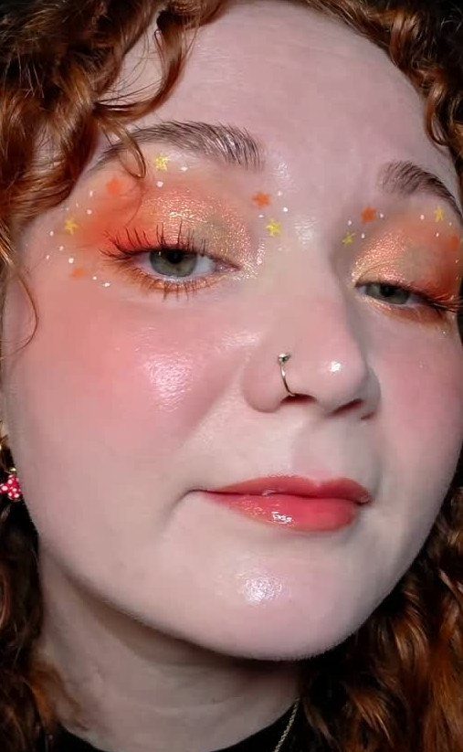 Person with orange glittery eyeshadow, star decals, and glossy coral-pink lips.