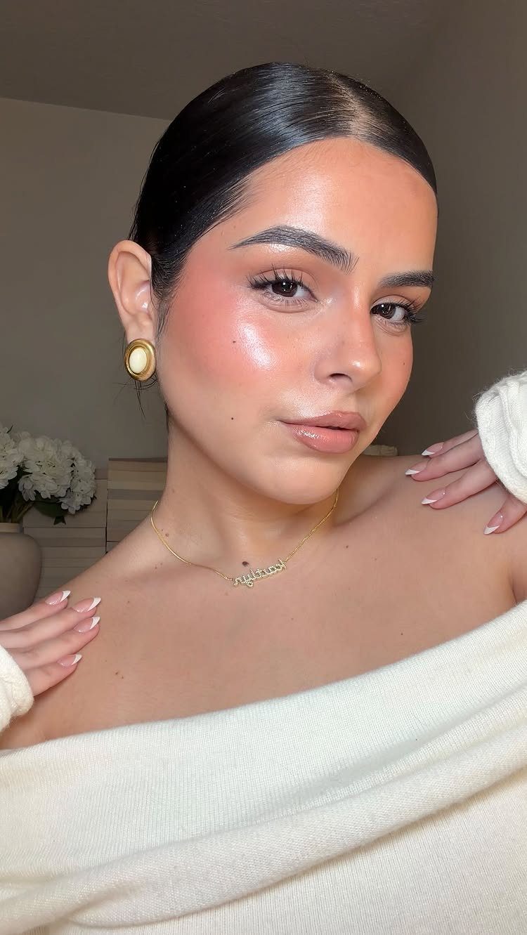 Woman with glowing skin, pink cream blush, and neutral makeup wearing white sweater.