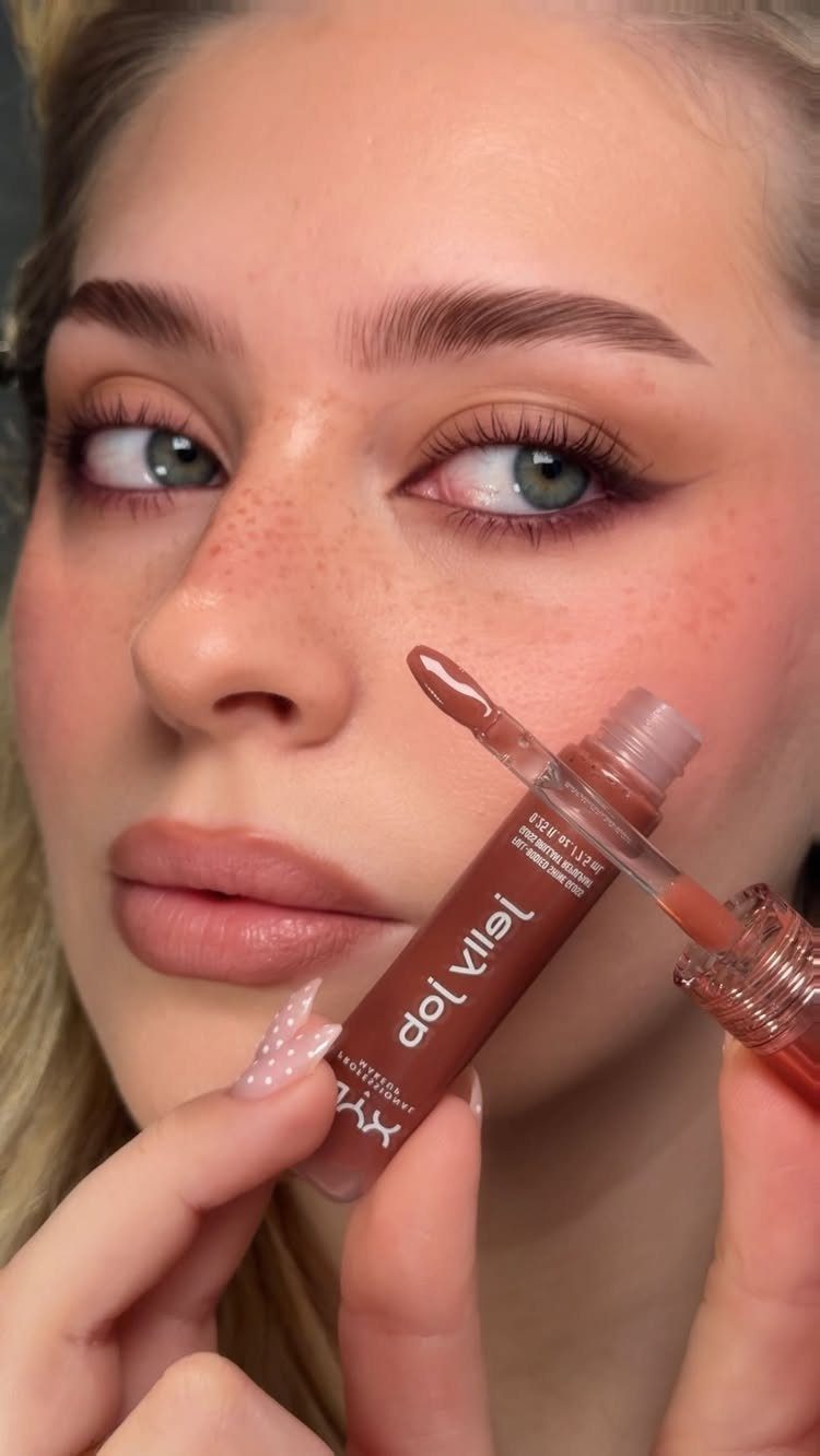 Woman with warm-toned makeup and freckles holding a brown lip gloss applicator.