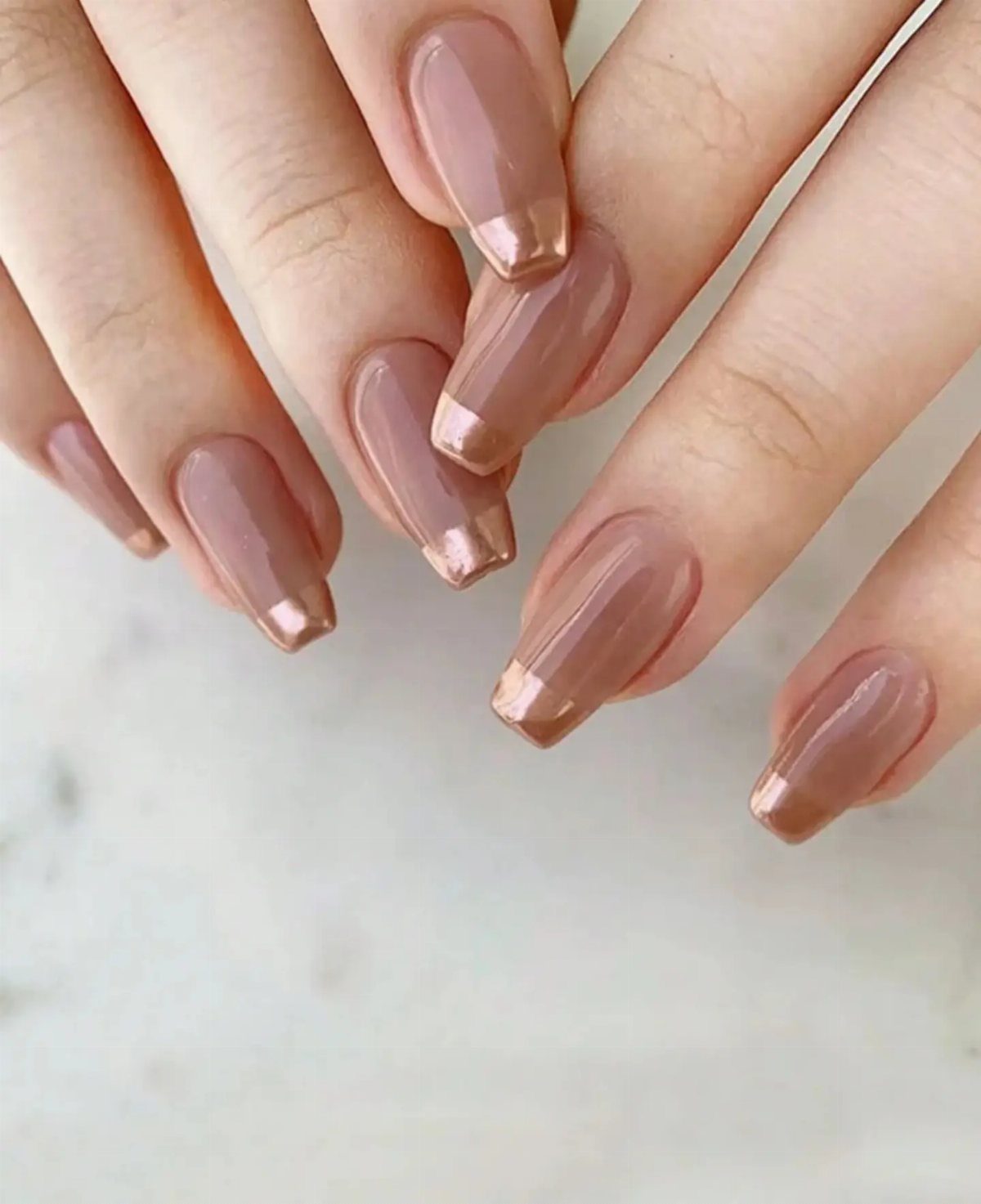Square nails with nude-pink polish and metallic gold tips on a marble surface.