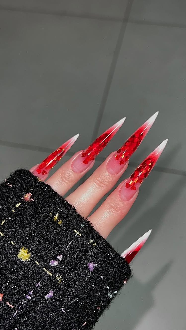 Stiletto nails with nude bases and red glitter tips featuring heart patterns.