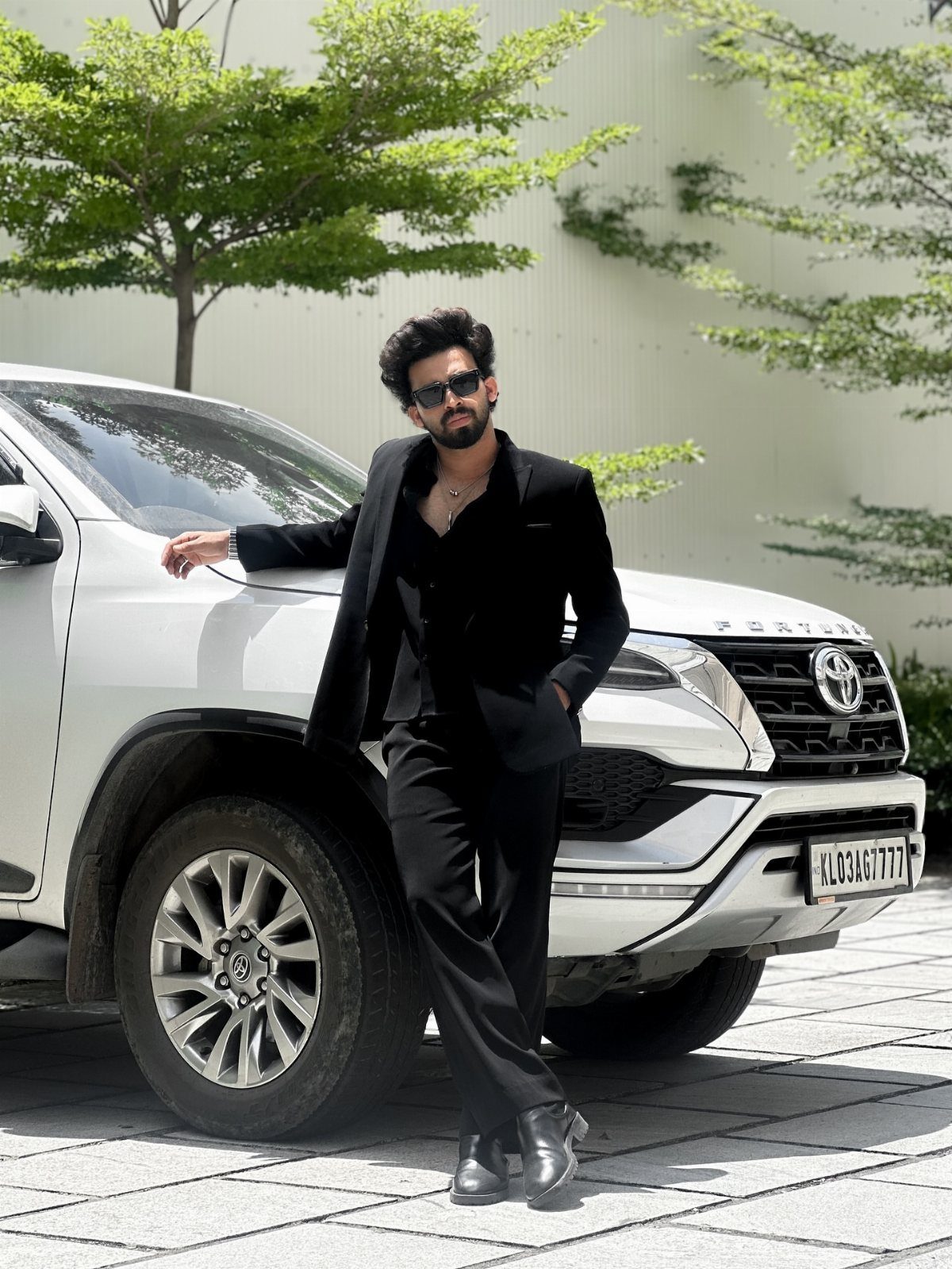 Man in a black suit and black shirt leaning against a white SUV.