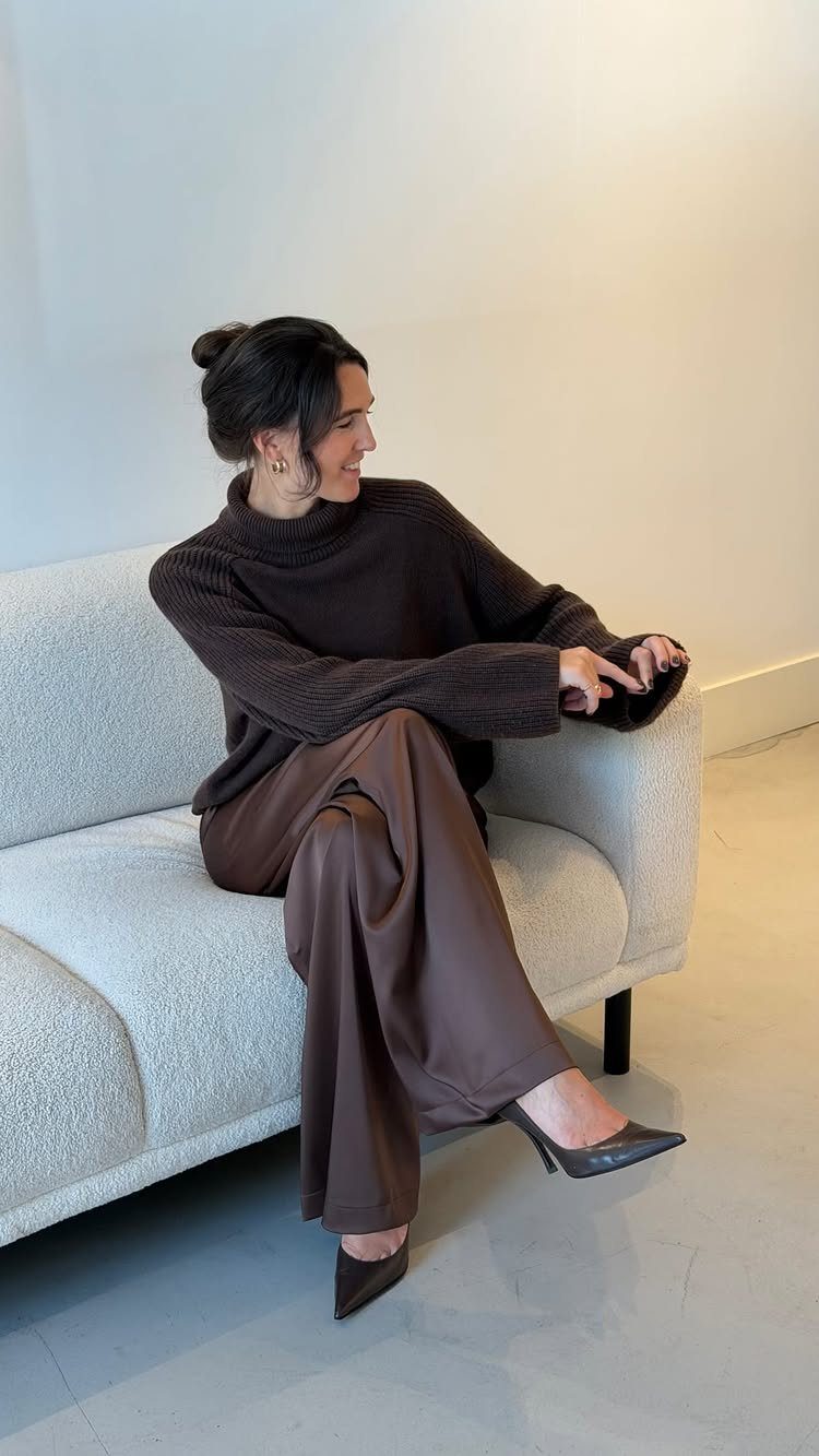 Woman wearing a brown oversized sweater and wide-leg satin trousers sitting on a sofa.