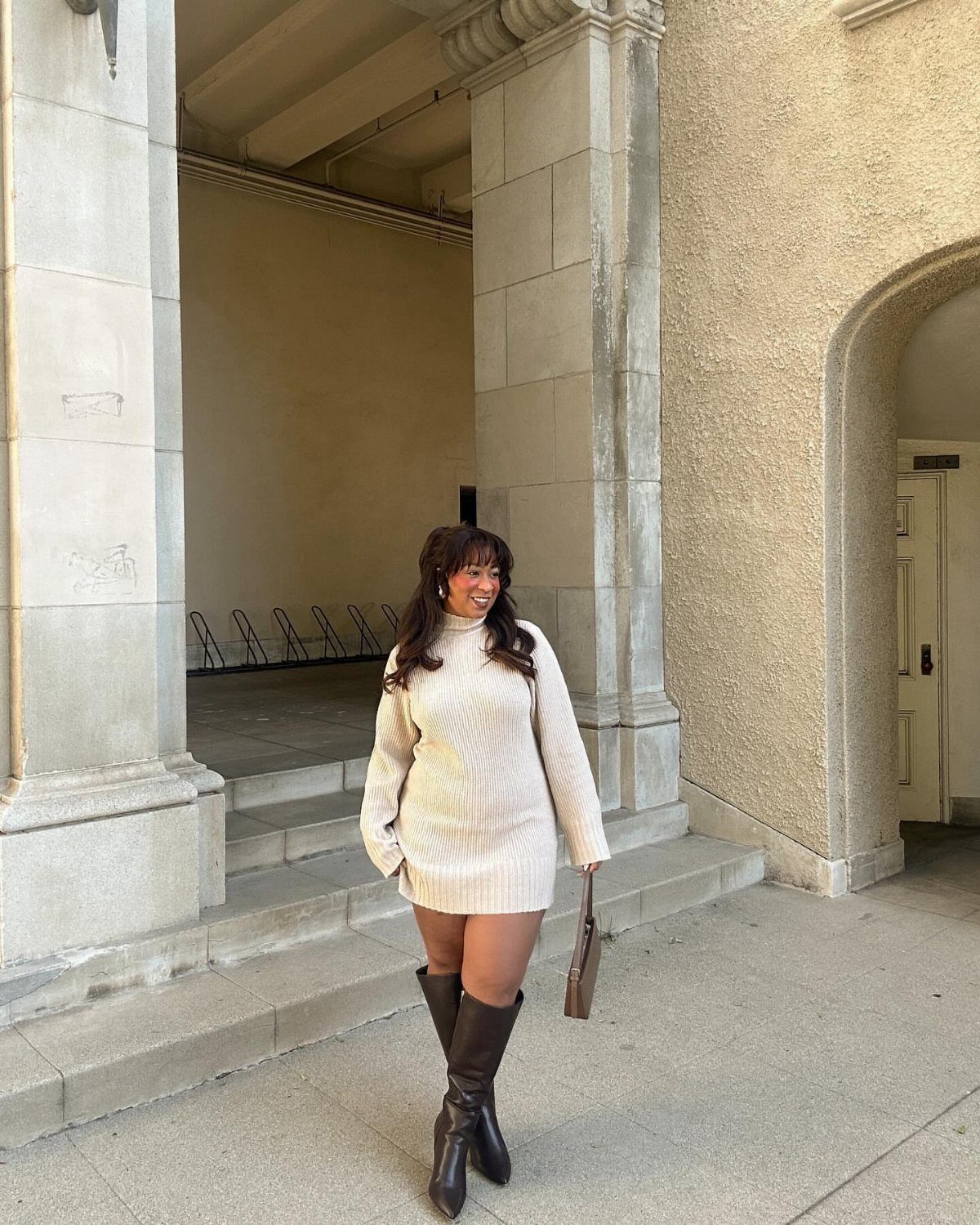 Woman wearing a cream knit sweater dress and tall brown leather boots outdoors.