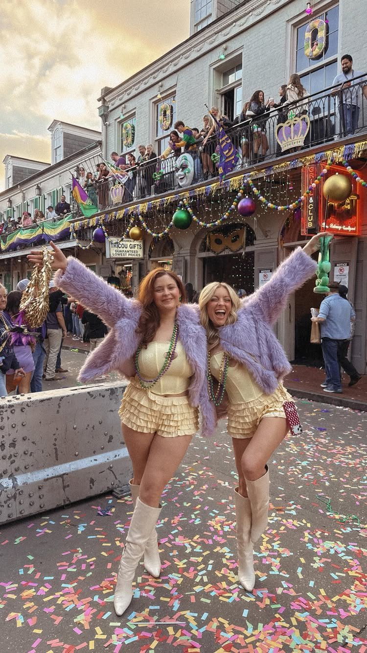 Two women in purple coats and yellow dresses posing on a confetti-covered street.