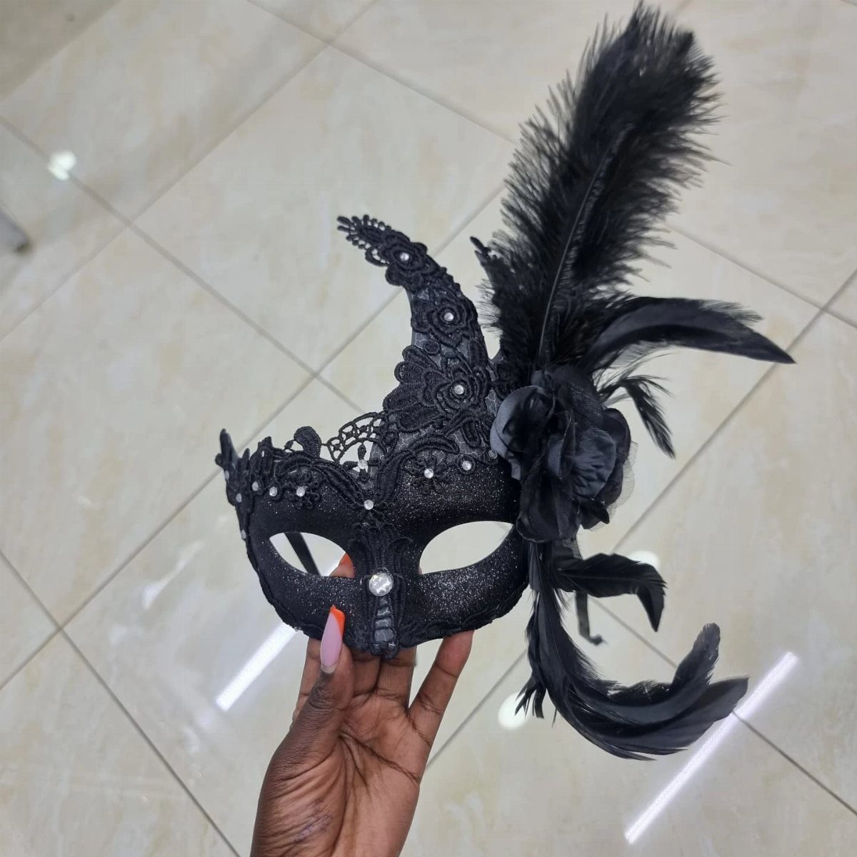 Ornate black masquerade mask with lace, sequins, and feathers held by a hand.