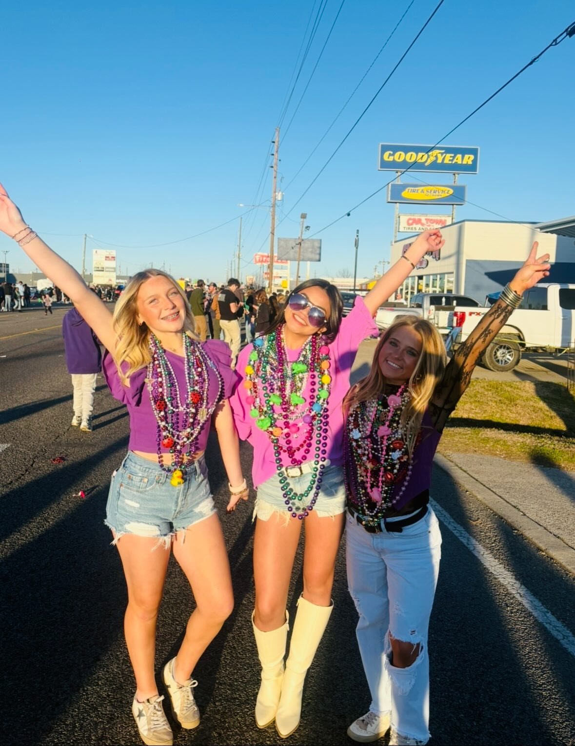 Three women in purple tops and denim wearing colorful beads on a street.