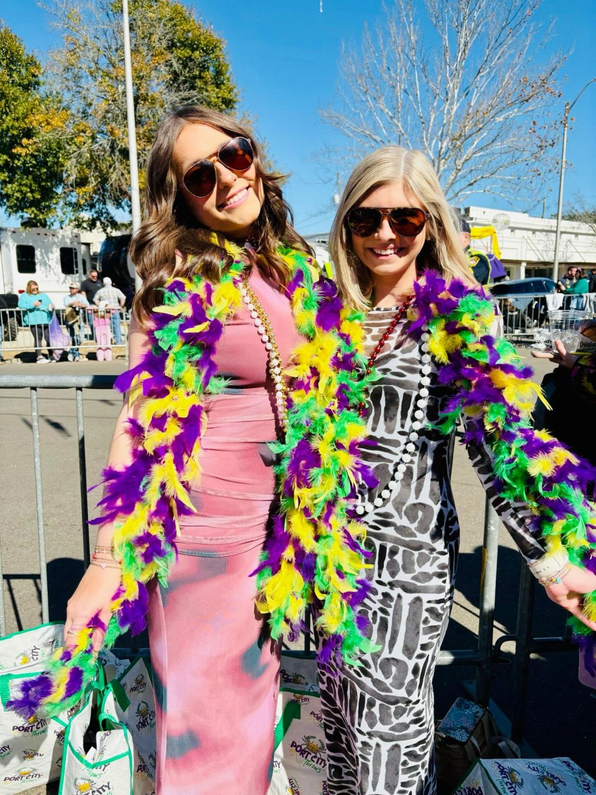 Two women wearing colorful Mardi Gras feather boas and sunglasses posing outdoors.