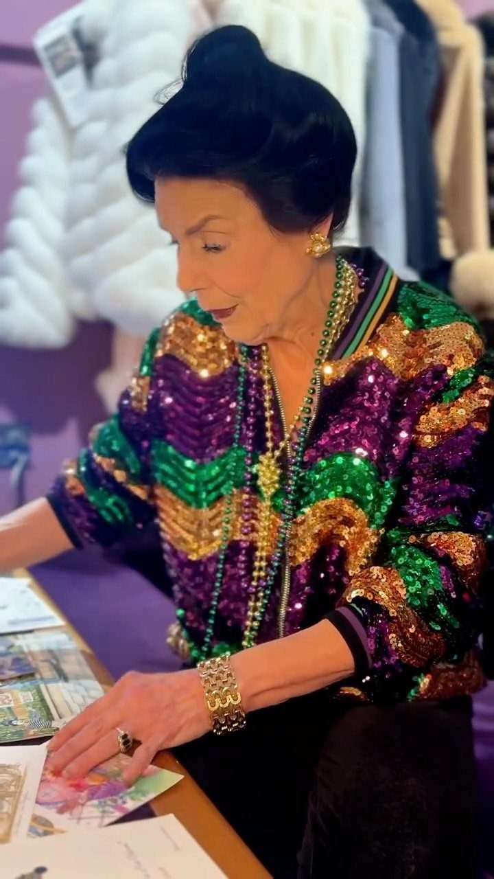 Woman wearing a purple, green, and gold sequined jacket and gold jewelry.
