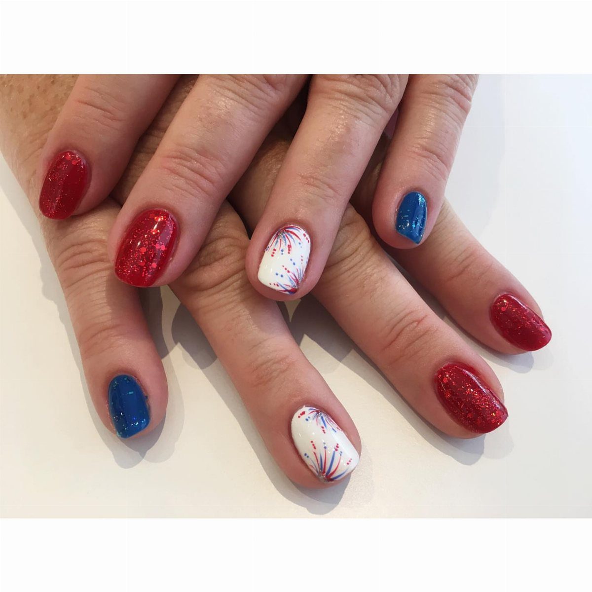 Manicured nails featuring red and blue glitter and white fireworks burst designs.