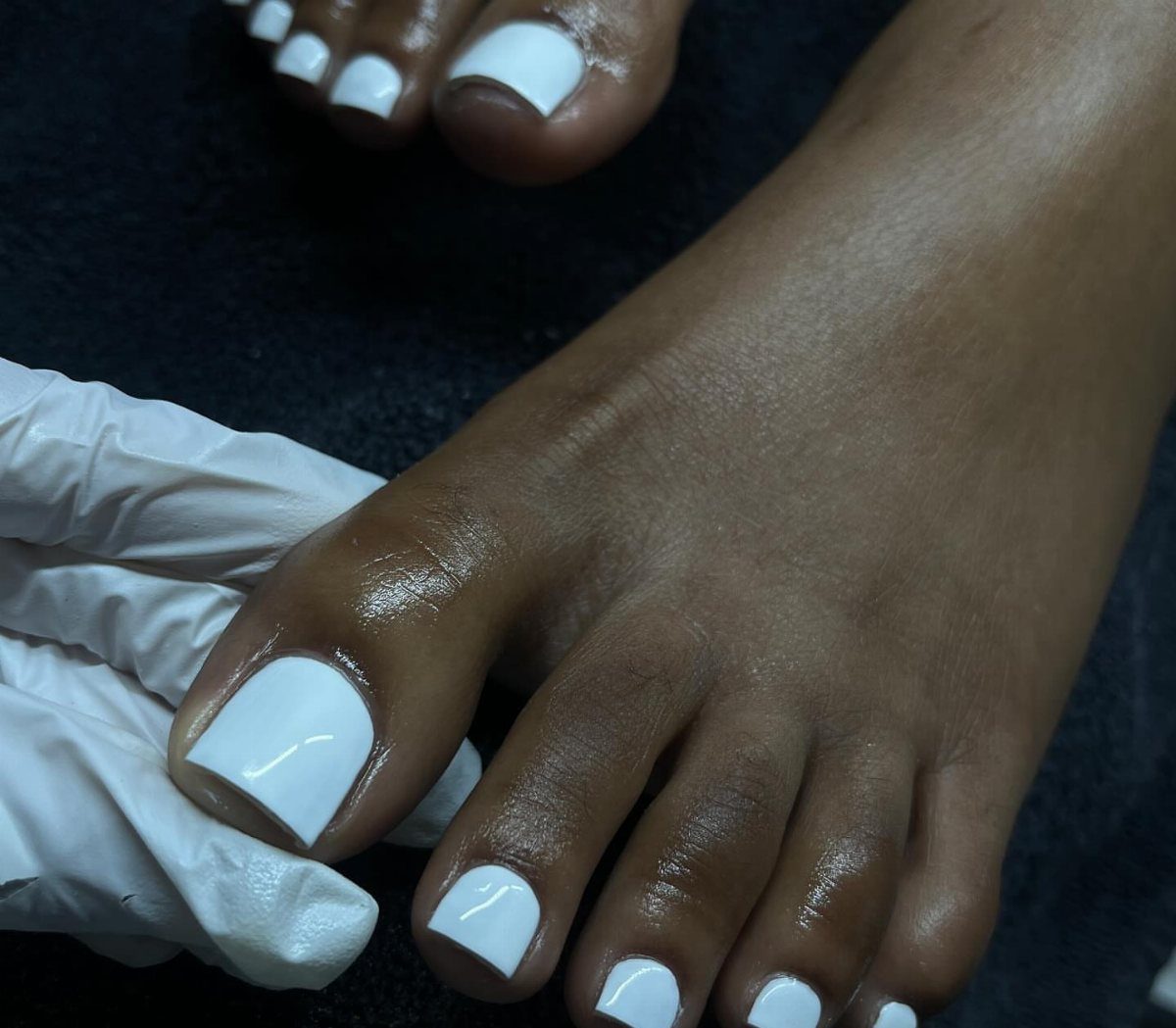Toes with solid white nail polish being held by a gloved hand.
