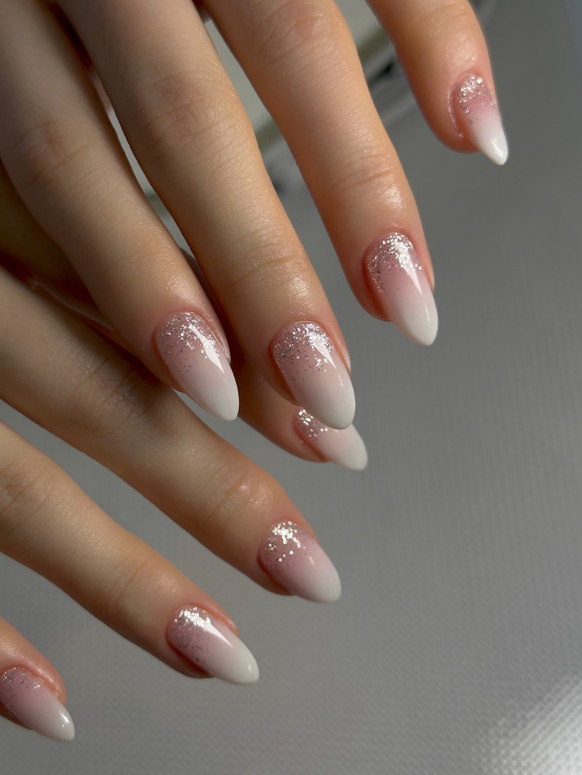 Almond-shaped nails with silver glitter gradient fading into white tips on a hand.